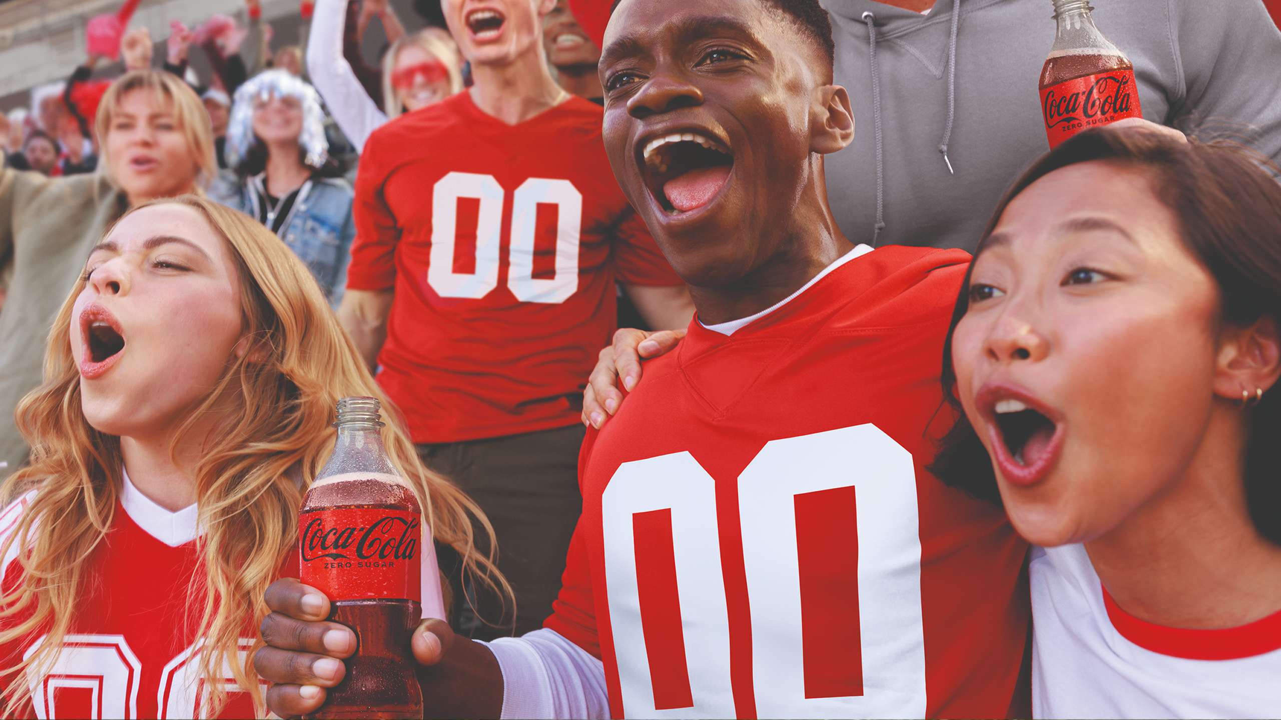 Excited football fan in red 00 jersey holding Coca-Cola Zero Sugar bottle while cheering in crowded stadium stands.