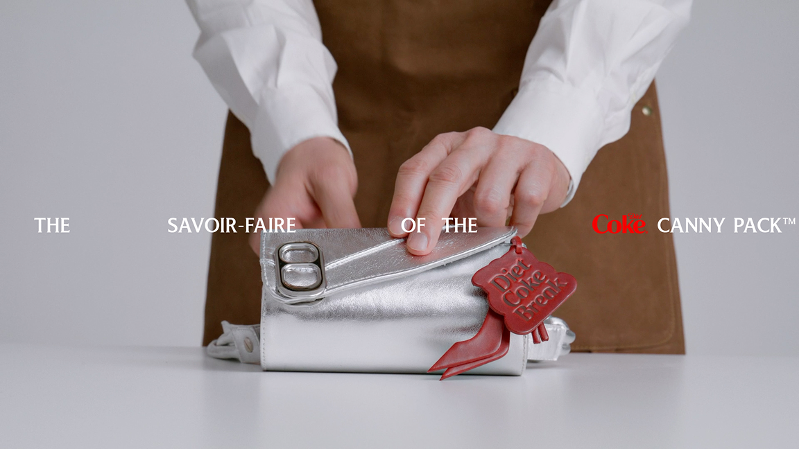 Hands in white shirt and apron presenting a silver Canny Pack bag with can-tab clasp and red Diet Coke Break tag on white table