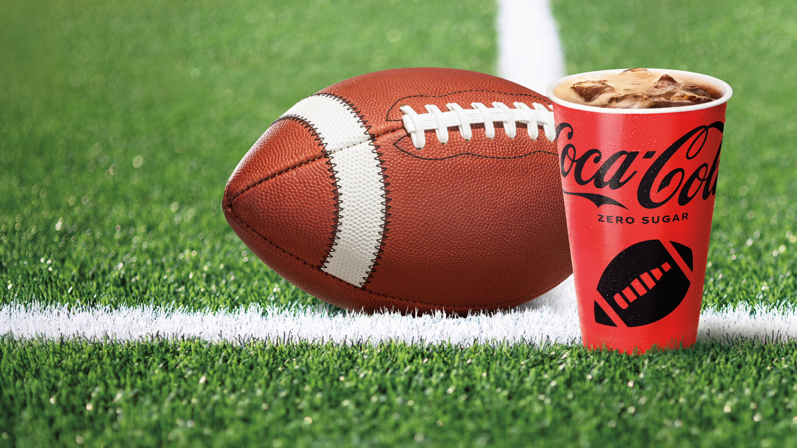 Football placed on a green field next to a Coca-Cola Zero Sugar cup with a football-themed design, perfect for fall football season promotions.