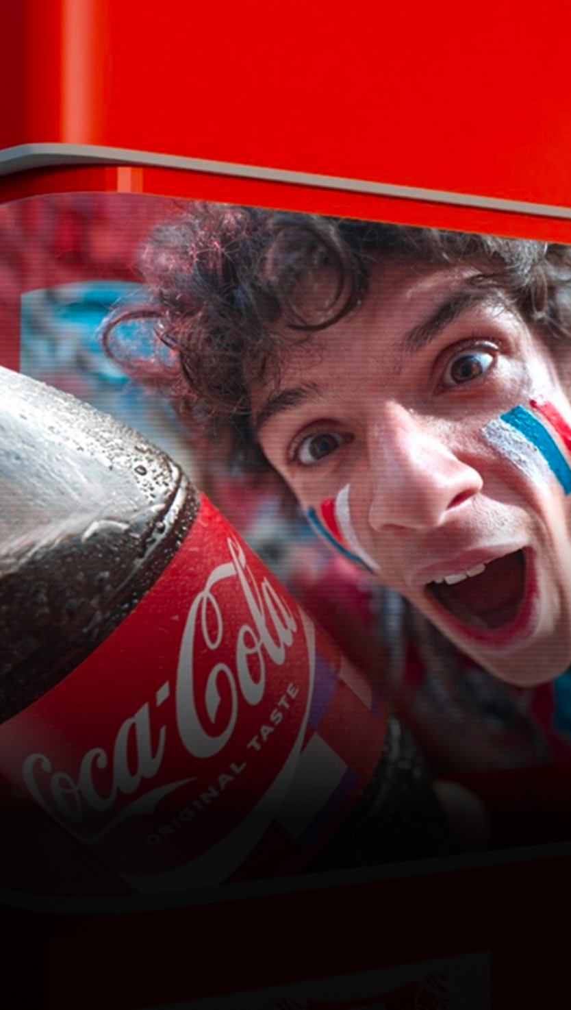 Close-up of chilled Coca-Cola bottle in ice with blurred cheering soccer fans in background at Coca-Cola Fan Zone.
