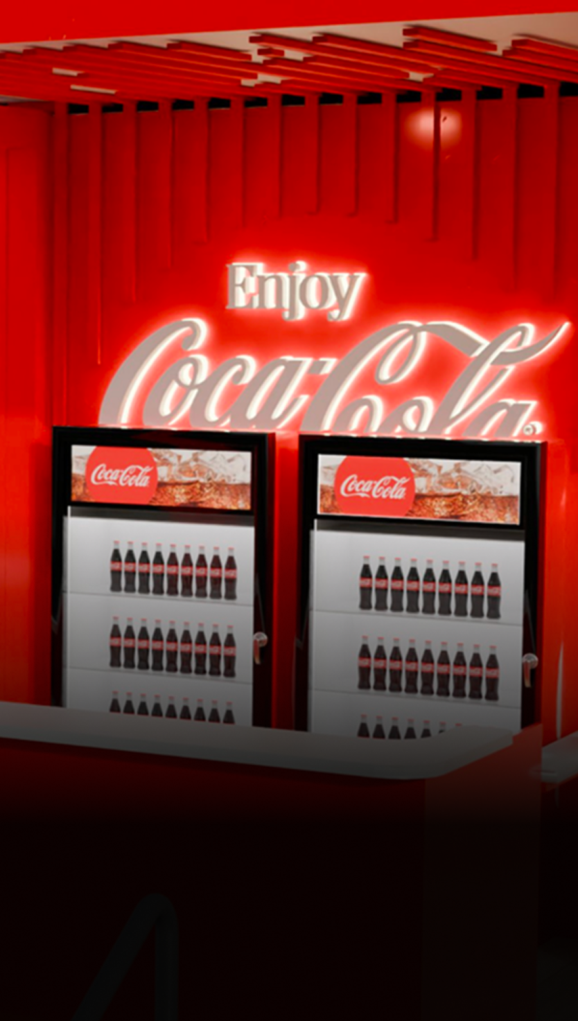 Enjoy Coca-Cola back bar with two coolers filled with contour bottles and illuminated red signage inside Fan Zone concession area.