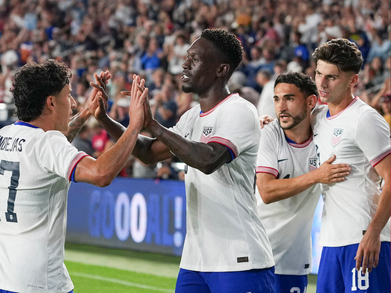 Group of United States men’s soccer players in white jerseys celebrating a goal together on the field in front of cheering FIFA World Cup fans.