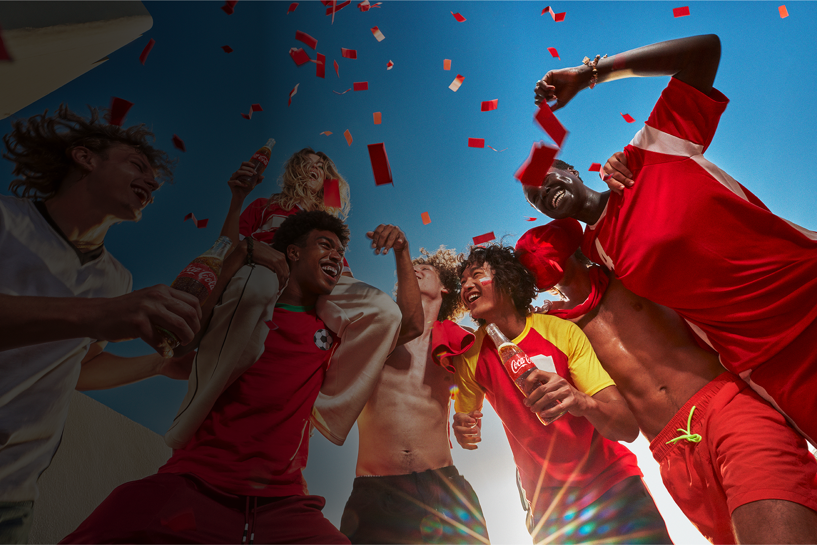 Group of young soccer fans in red jerseys celebrating under falling confetti, raising Coca‑Cola bottles against blue sky