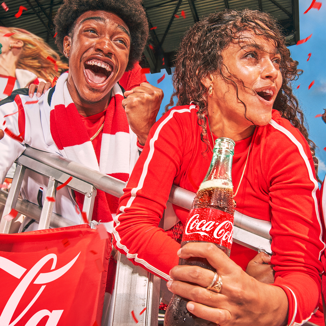 Two cheering fans hold a Coca-Cola bottle in stadium seating with falling confetti for Soccer Central.