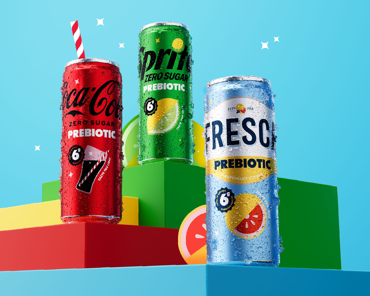 Three condensation-covered prebiotic soda cans displayed on colorful geometric blocks against a light blue background: Coca-Cola Zero Sugar Prebiotic (with a red-and-white straw), Sprite Zero Sugar Prebiotic, and Fresca Prebiotic Grapefruit Citrus, each marked with a "6g fiber to support digestive health" badge.