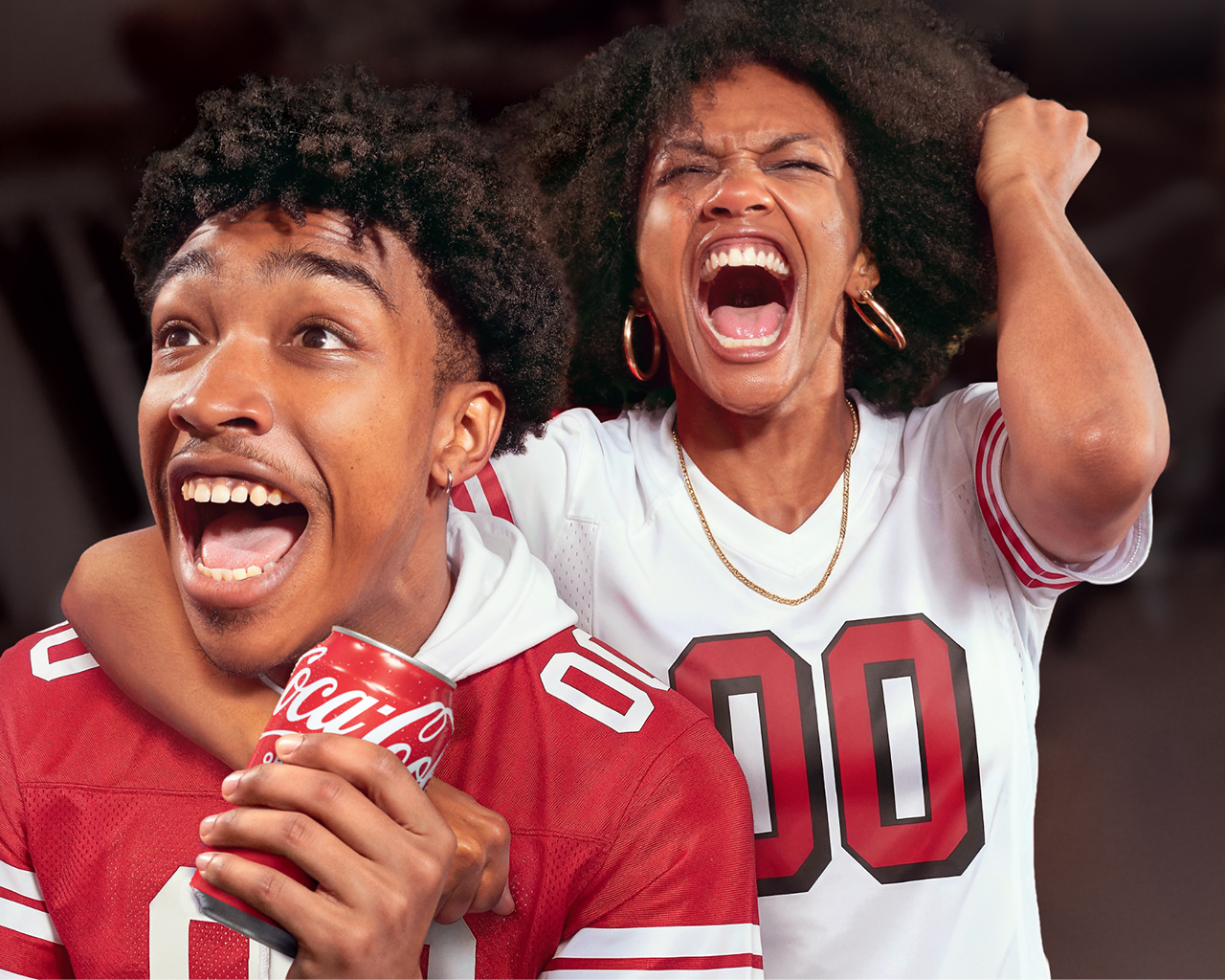 A young man and woman wearing white and red football jerseys with the number "00" cheer excitedly, mouths open in celebration; the man holds a Coca-Cola can with the logo visible.