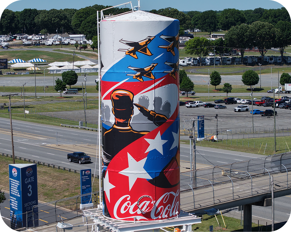 Acercamiento del mural de Coca‑Cola de Amber Thompson en una torre de agua, mostrando aviones y una figura saludando.