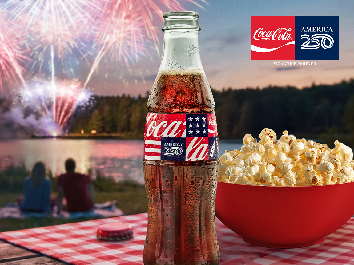 Botella de vidrio Coca‑Cola America 250 sobre mesa de picnic con palomitas mientras pareja mira fuegos artificiales.