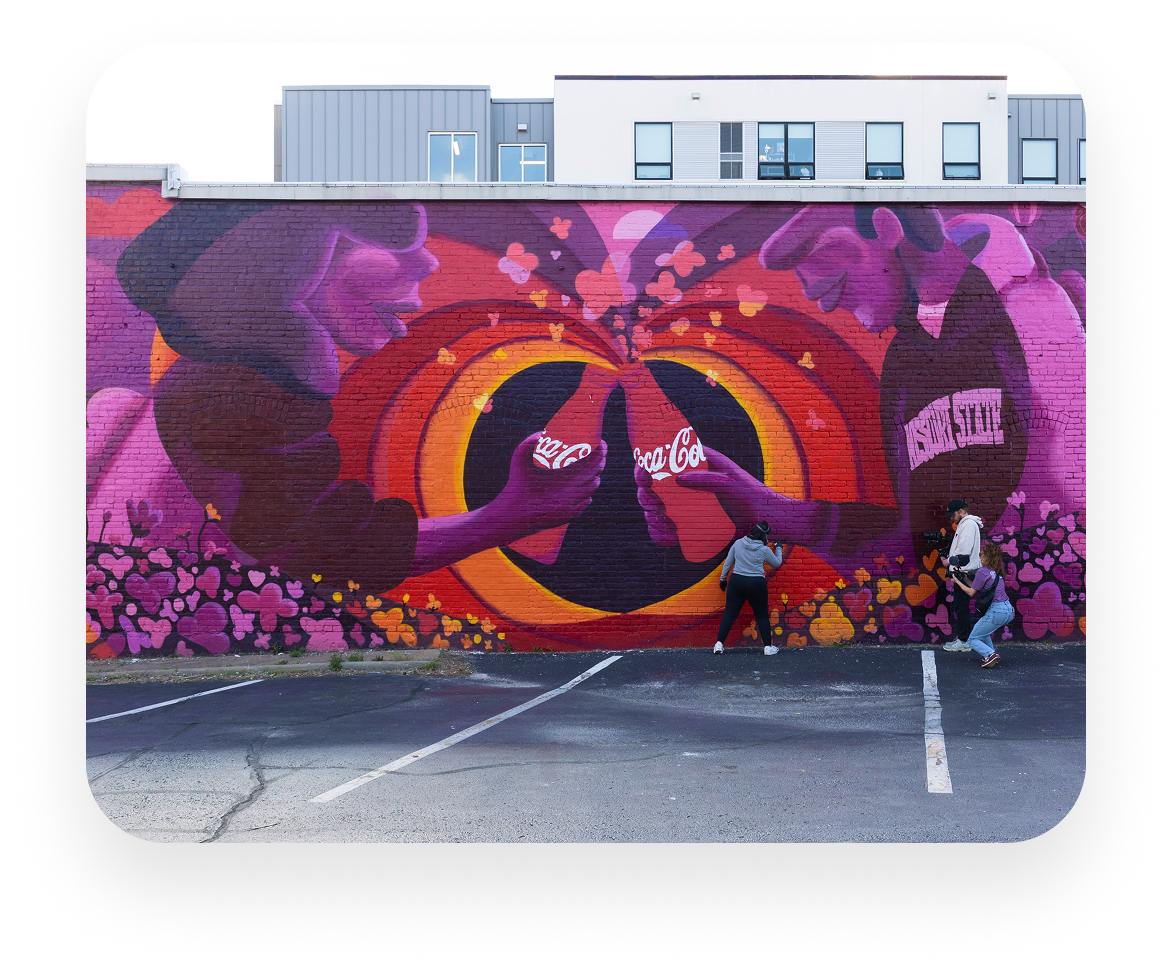 La artista Meg Wagler sosteniendo una lata de pintura en aerosol frente a su vibrante mural de Coca‑Cola.