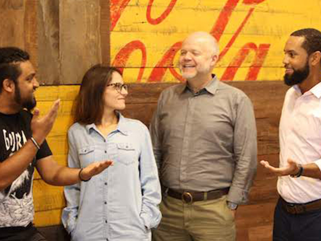 Um grupo de quatro pessoas conversa e sorri em frente a uma parede de madeira com a logomarca da Coca-Cola